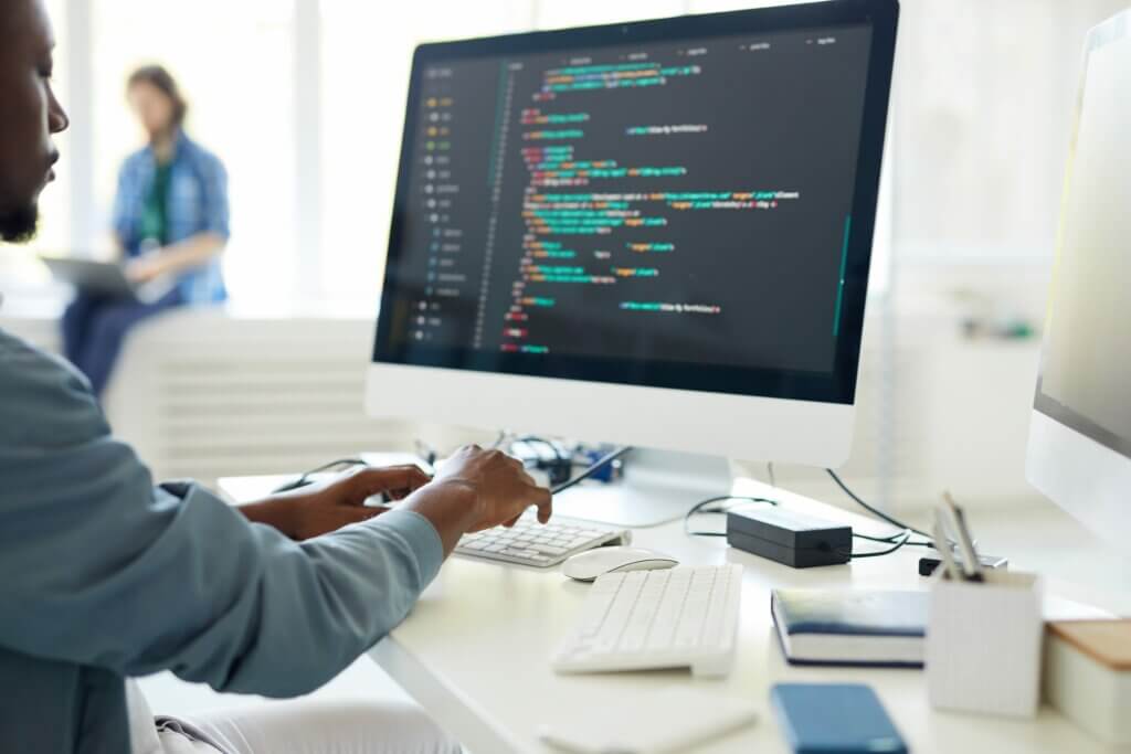 Programas de formação Zup: Homem negro programando em uma mesa branca com agenda e celular. Ao fundo, uma mulher mexendo no notebook.